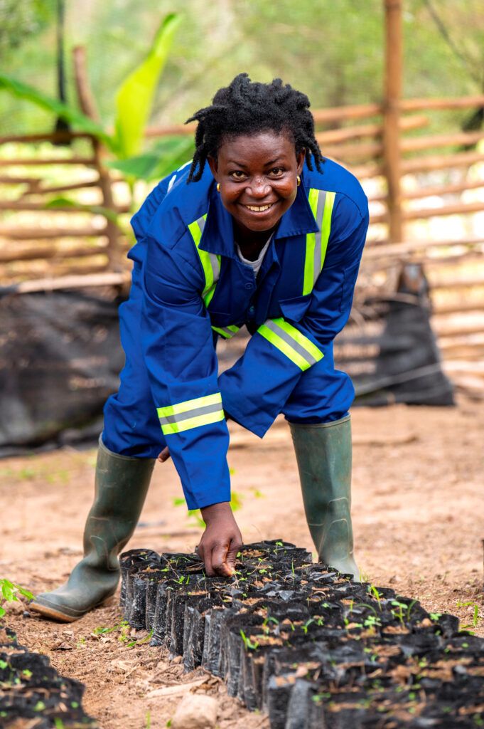 a woman smiles as she's crouching over tree seedlings on the ground